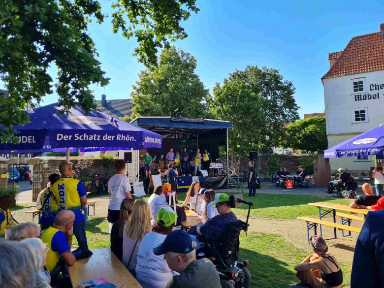 Einige Menschen sitzen vor einer Bühne in einem großen Garten.