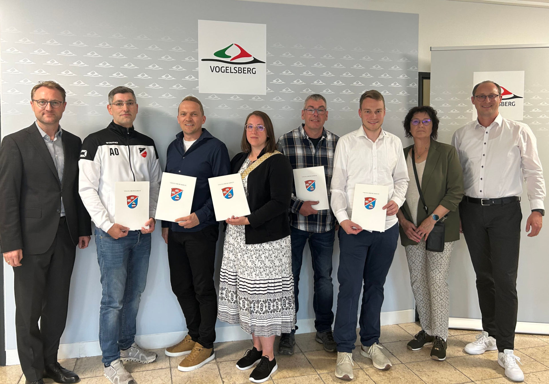 Acht Personen haben sich vor einer Fotowand zum Gruppenbild aufgestellt. Sie präsentieren Förderbescheide.