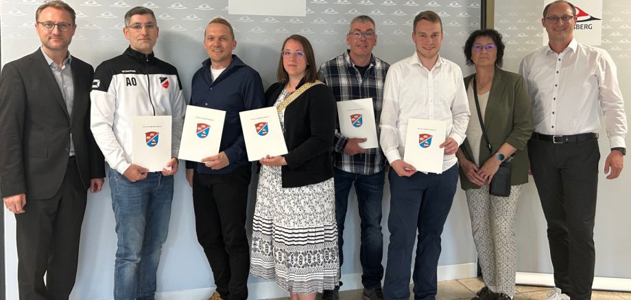 Acht Personen haben sich vor einer Fotowand zum Gruppenbild aufgestellt. Sie präsentieren Förderbescheide.