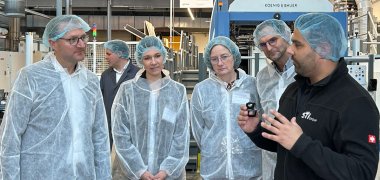 Mehrere Personen stehen - bekleidet mit Hygieneschutzanzügen - in einer Druckerei und sprechen miteinander.