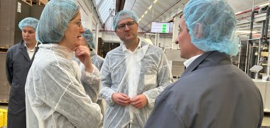 Drei Personen stehen - bekleidet mit Hygieneschutzanzügen - in einer Druckerei und sprechen miteinander.