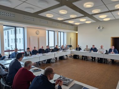 Eine Gruppe von Menschen sitzt in einem Sitzungssaal an Tischen. Sie sind im Gespräch.