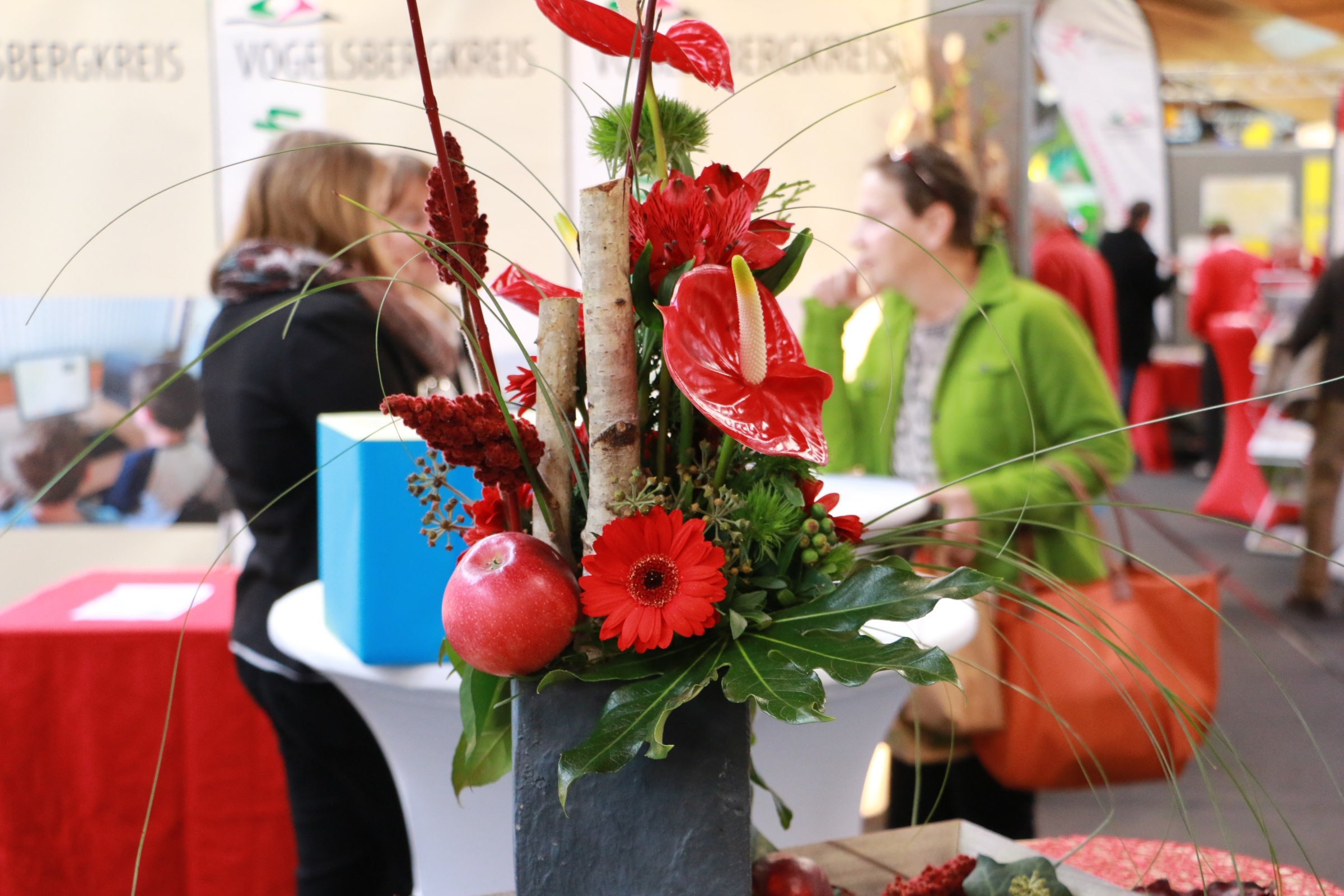 Im Vordergrund ist ein Gesteck mit roten Blumen, Holz und allerlei Grün zu sehen. im Hintergrund sprechen drei Menschen miteinander vor einem Messestand.