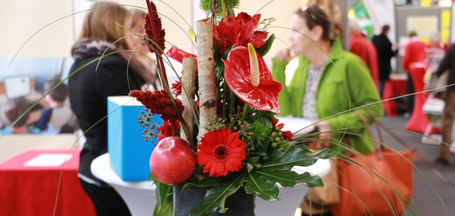 Im Vordergrund ist ein Gesteck mit roten Blumen, Holz und allerlei Grün zu sehen. im Hintergrund sprechen drei Menschen miteinander vor einem Messestand.
