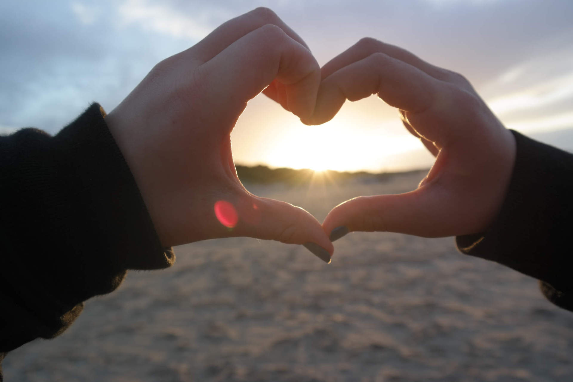Zwei Hände formen ein Herz. im Hintergrund ist ein Sonnenuntergang am Strand zu sehen