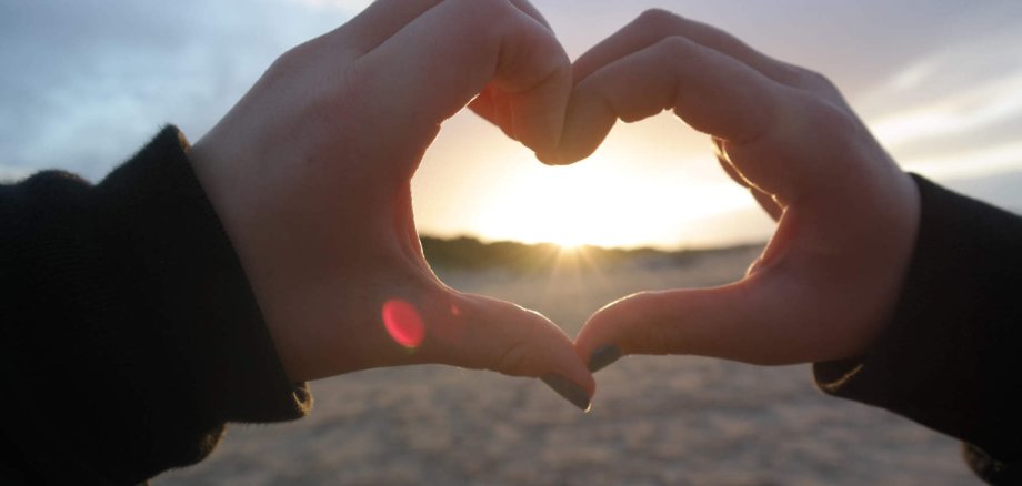 Zwei Hände formen ein Herz. im Hintergrund ist ein Sonnenuntergang am Strand zu sehen