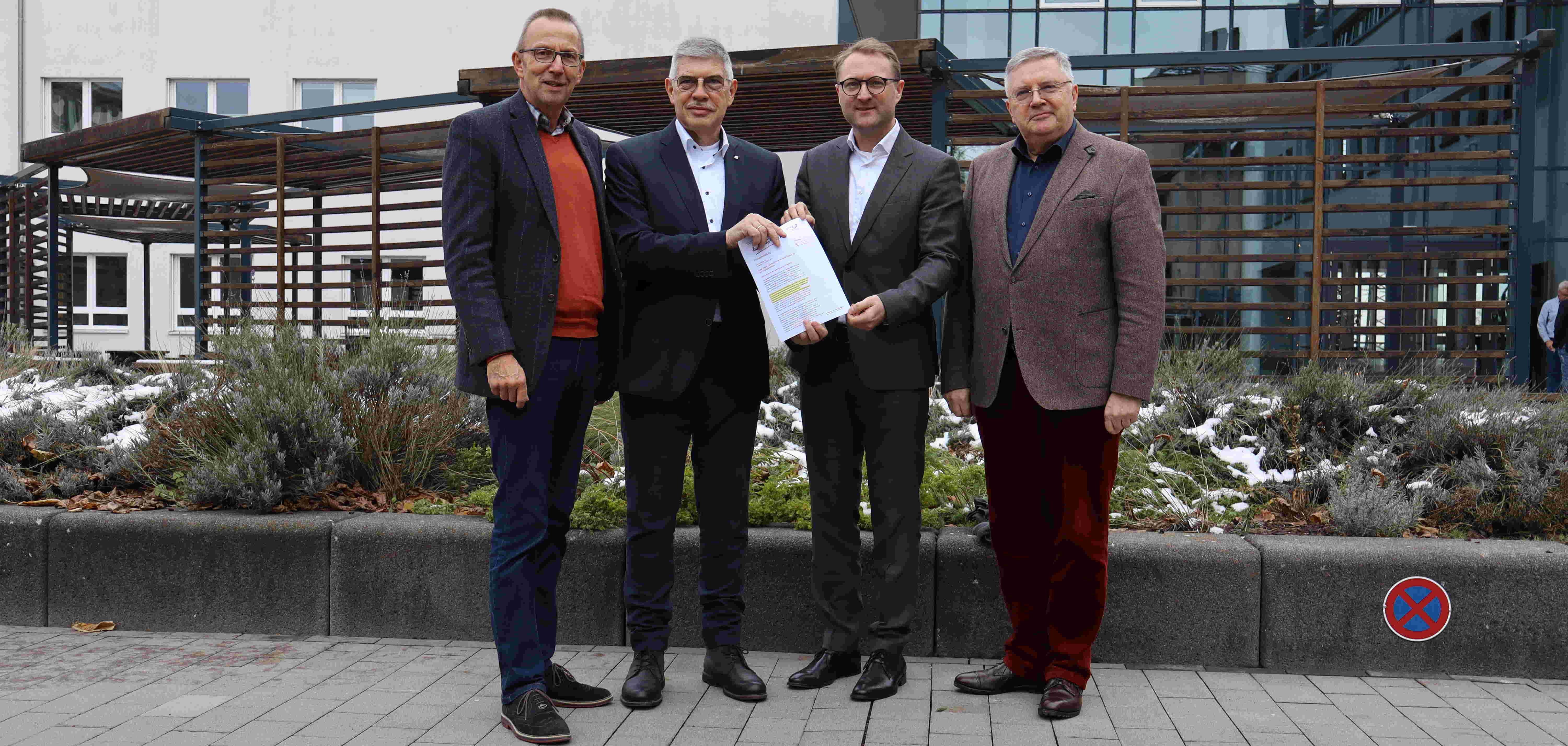 Die beiden OVAG-Vorstände Joachim Arnold (links) und Oswin Veith (rechts) mit dem Landrat des Vogelsbergkreises, Manfred Görig (Zweiter von links) und dem Ersten Kreisbeigeordneten Dr. Jens Mischak. Sie stehen vor der OVAG-Verwaltung und präsentieren das Konzept in einer Mappe. 