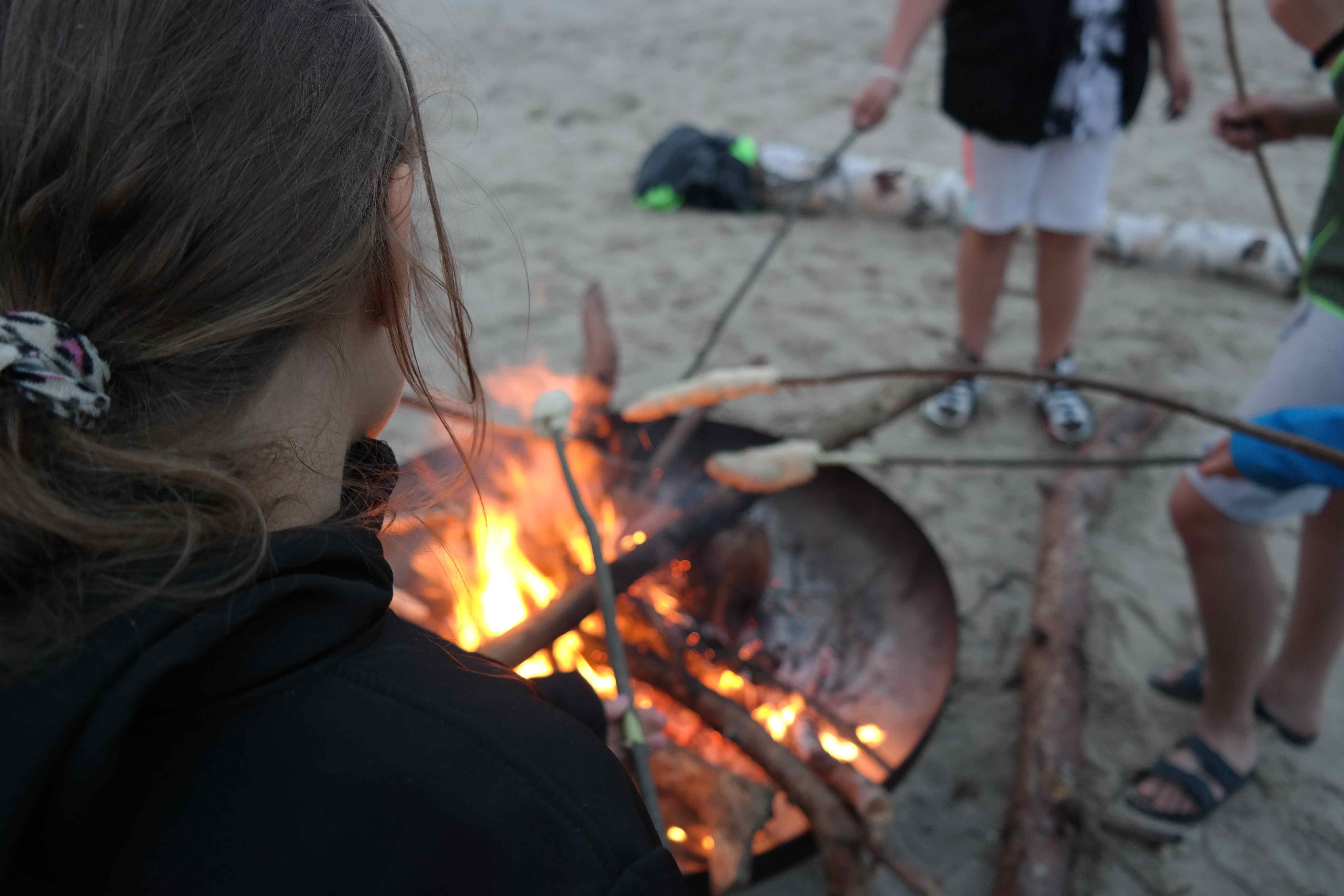 Jugendliche backen über einer Feuerschale Stockbrot. Es sind keine Gesichter zu sehen, das Foto ist über eine Schulter hinweg aufgenommen.
