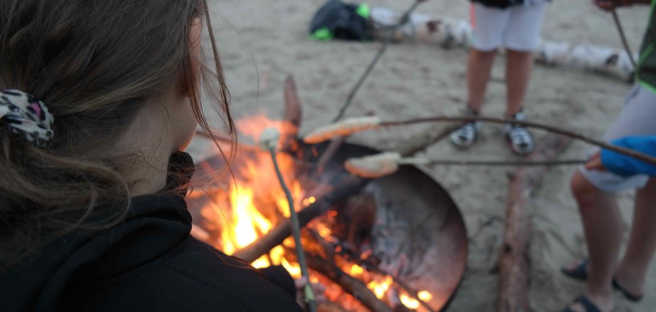 Jugendliche backen über einer Feuerschale Stockbrot. Es sind keine Gesichter zu sehen, das Foto ist über eine Schulter hinweg aufgenommen.