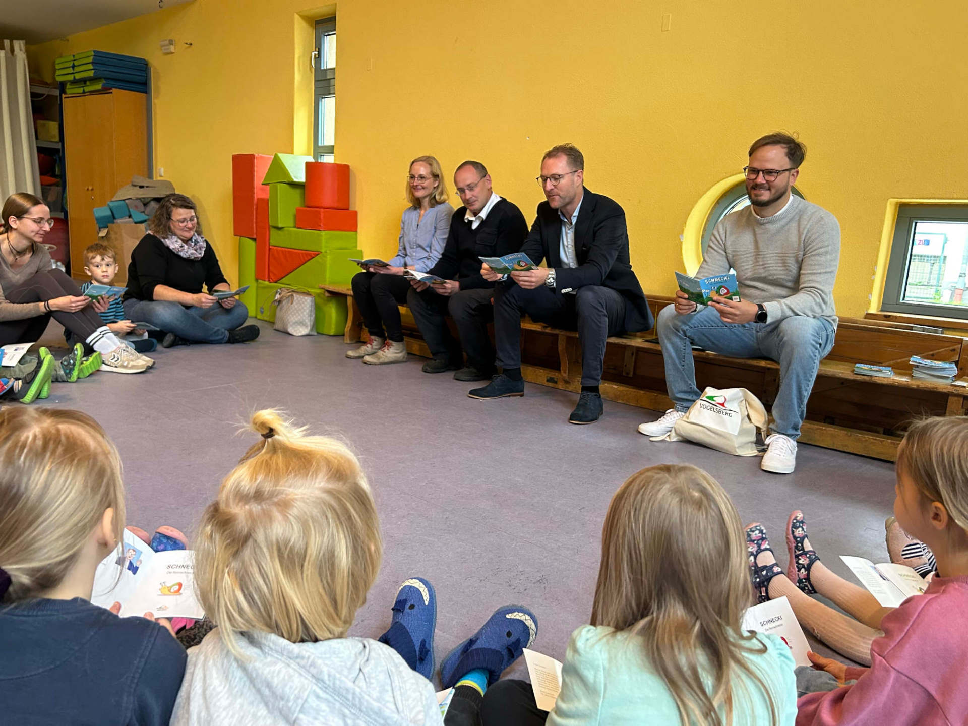 vier Erwachsene sitzen in einem Kindergarten au feiner Bank und lesen einigen Kindern aus einem Kinderbuch vor.