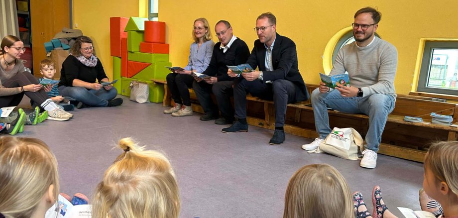 vier Erwachsene sitzen in einem Kindergarten au feiner Bank und lesen einigen Kindern aus einem Kinderbuch vor.
