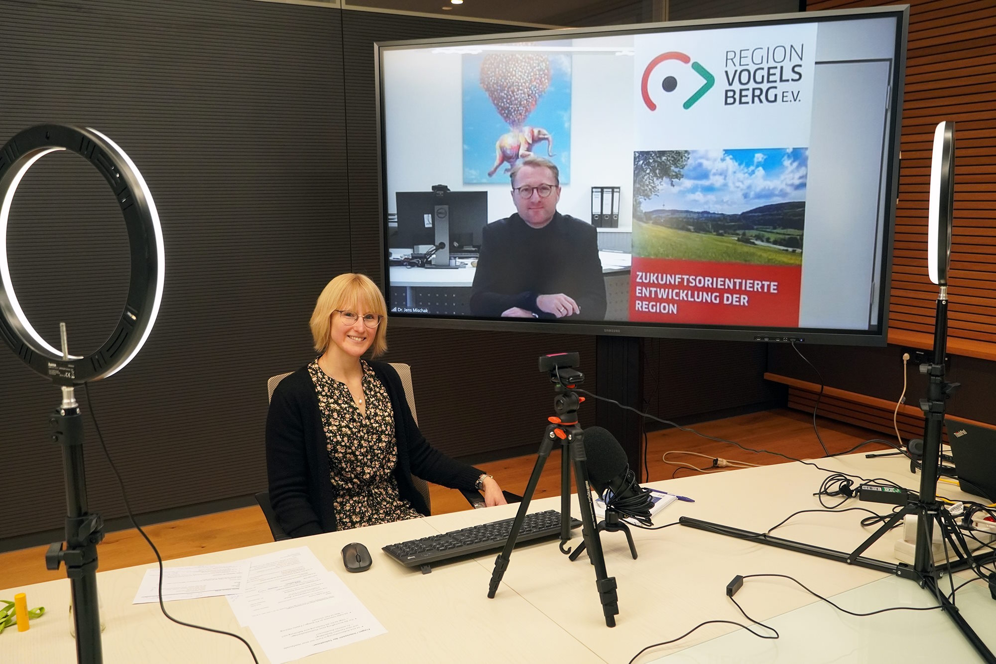 Dr. Jens Mischak, Erster Kreisbeigeordneter des Vogelsbergkreises und Vorstandsvorsitzender des Vereins Region Vogelsberg, gemeinsam mit Johanna Dunez, Kundenberaterin Energiedienstleistungen der OVAG, während des Online-Seminars. Dr. Mischak ist auf einem Bildschirm zu sehen, Frau Dunez sitzt vor Kameras und Beleuchtung. 