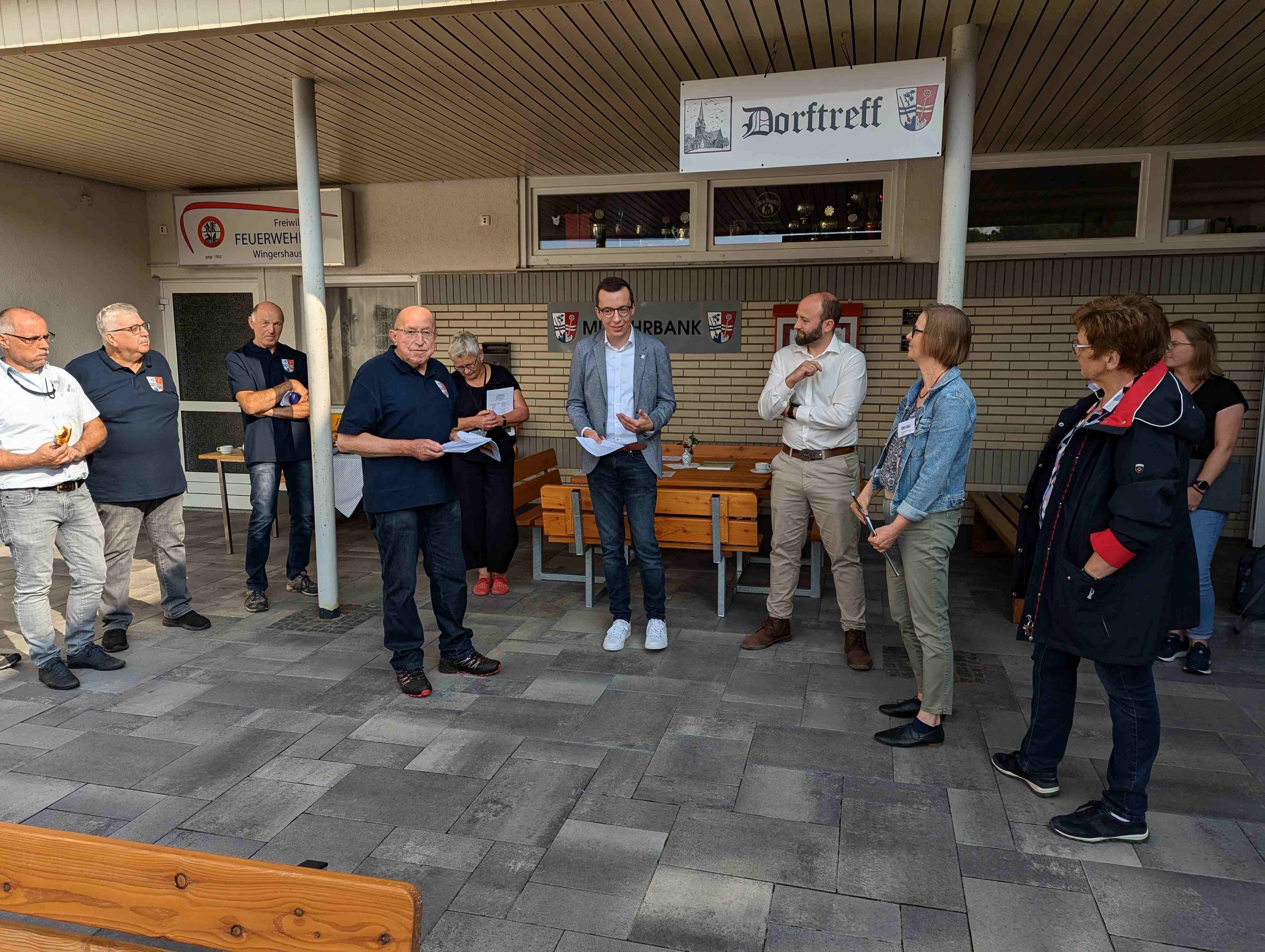 Mehrere Menschen stehen vor ein paar Sitzgelegenheiten und halten eine Ansprache. Patrick Krug, Erster Kreisbeigeordneter, eröffnet einen WEttbewerb unter Dörfern
