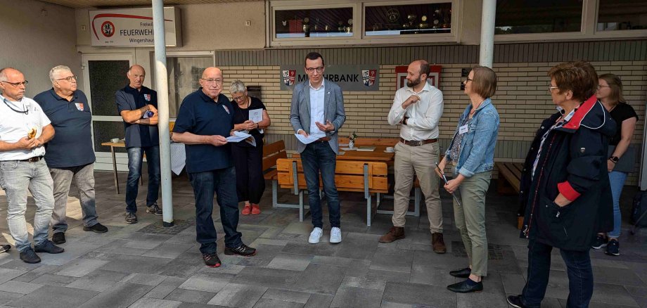 Mehrere Menschen stehen vor ein paar Sitzgelegenheiten und halten eine Ansprache. Patrick Krug, Erster Kreisbeigeordneter, eröffnet einen WEttbewerb unter Dörfern