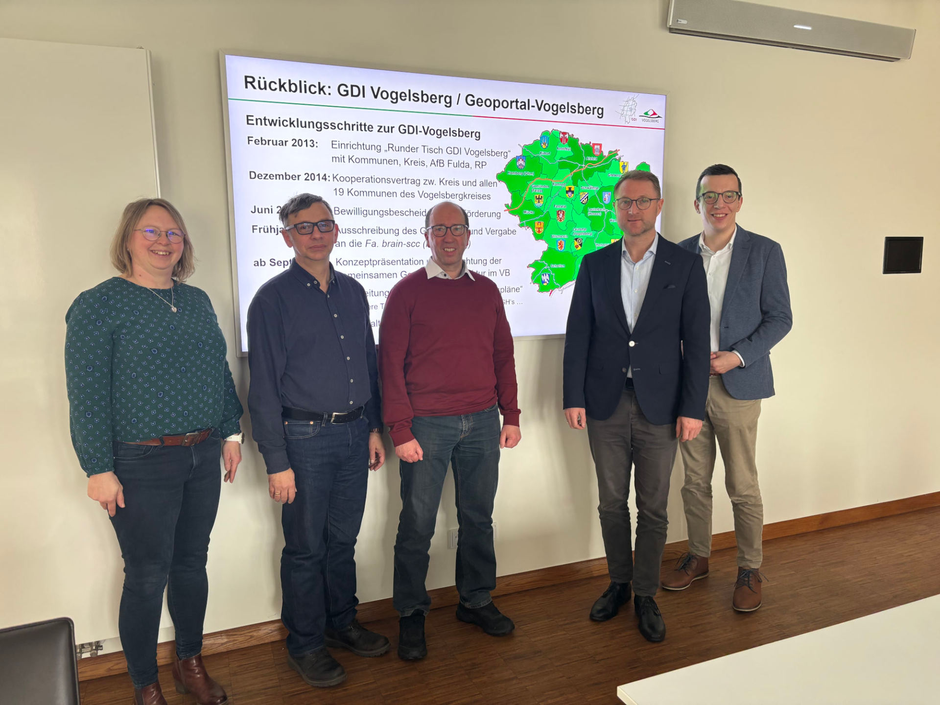 Fünf Personen stehen vor einem Präsentationsbildschirm und lächeln in die Kamera